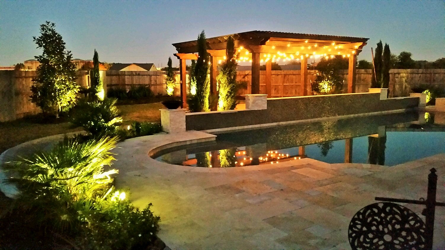 Nighttime backyard with illuminated pool, pergola with string lights, and landscaping.
