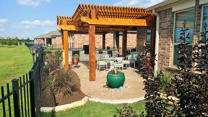 Patio with pergola, outdoor kitchen, seating, and fountain in a yard. Brick house in the background.