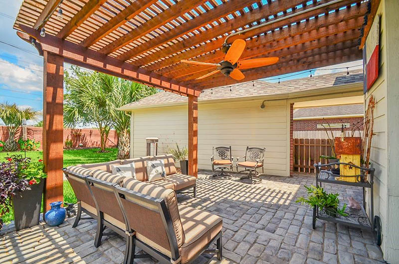 Patio with pergola, seating, brick flooring, and a ceiling fan; lush green grass and a house in the background.