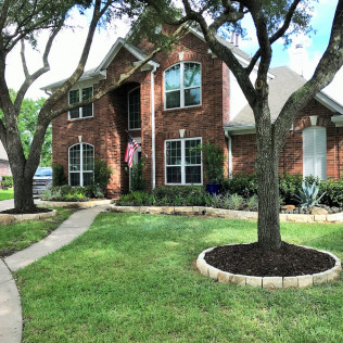 Two-story brick house with manicured lawn, landscaping, and shade trees. A flag is in the center.