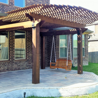 Wooden pergola with swing, over a stone patio, next to a brick house.