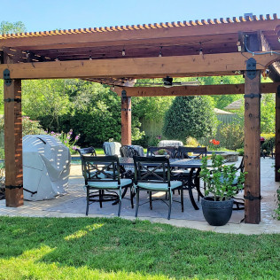 Pergola over outdoor dining table with chairs, surrounded by greenery and lawn.