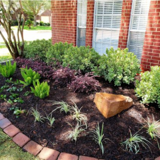 Landscaped flower bed with red brick border, featuring green, purple, and light green plants against a red brick wall.
