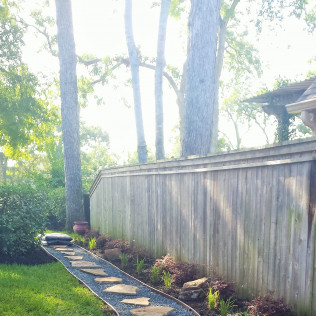 Stone pathway leads beside a wooden fence through a yard with trees and flowers.