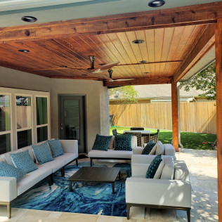 Outdoor patio with white sofas, blue rug, wooden ceiling, and backyard view.