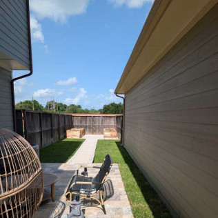 Patio with furniture, grass, and raised garden beds along a wooden fence on a sunny day.