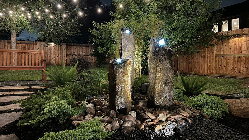Backyard fountain with three stone columns, illuminated at night. String lights, lush greenery, wooden fence.