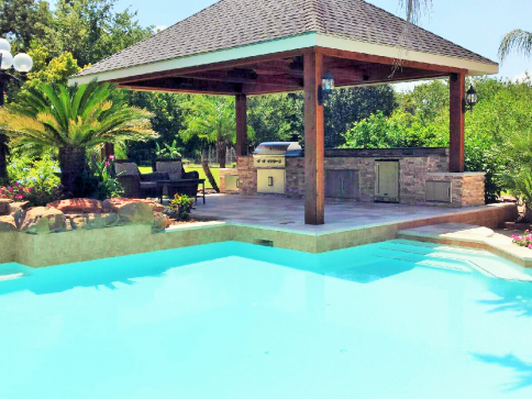 Poolside outdoor kitchen with a gazebo, grill, and lush landscaping.