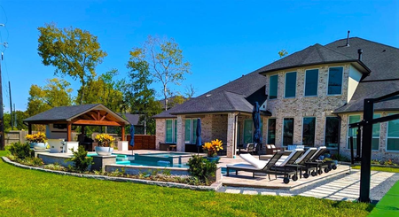 Backyard with pool, outdoor kitchen, and two-story brick house under a blue sky.