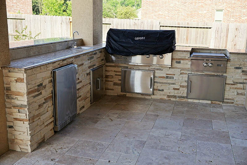 Outdoor kitchen with stone veneer and stainless steel appliances. Sink, grill, and storage cabinets visible.