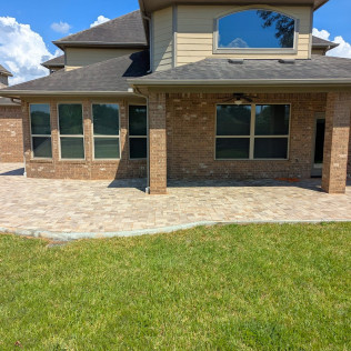 Backyard patio with brick columns, brick wall, and a curved stone border. Green lawn and a two-story beige house in the background.