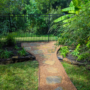 Stone path with gravel border leads to black metal fence in a garden.