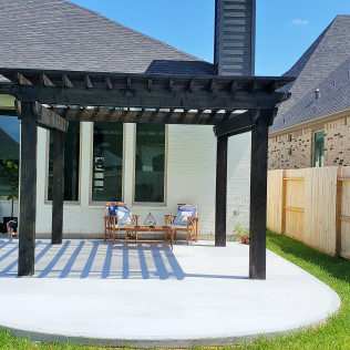 Black pergola over a gray concrete patio with two chairs and a small table.