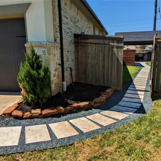 Stone path leads to a wooden fence next to a building with a small bush and flower bed.
