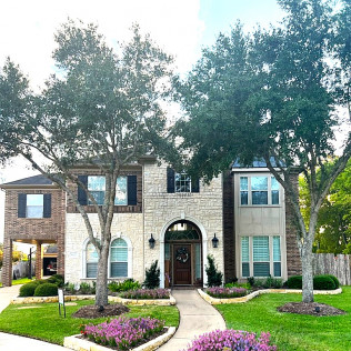Two-story house with stone and brick facade, front walkway, manicured lawn, and flowering beds.