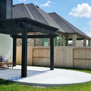 Black pergola on a concrete patio next to a wooden fence and house. Blue sky.