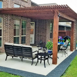 Patio with wooden pergola, seating, and landscaping next to a brick house.