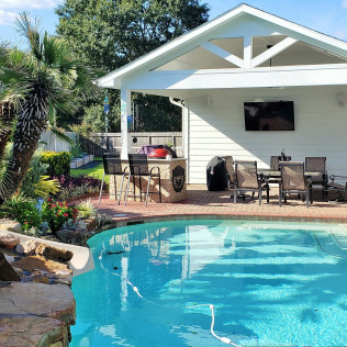 Poolside patio with bar, TV, and dining area under a white roof. Palm trees and blue sky visible.