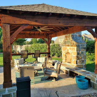 Wooden gazebo with fireplace, outdoor seating, and lush greenery.
