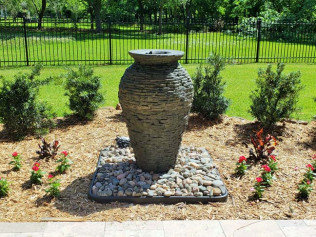 Stone urn water fountain in a garden bed with landscaping and a black fence backdrop.