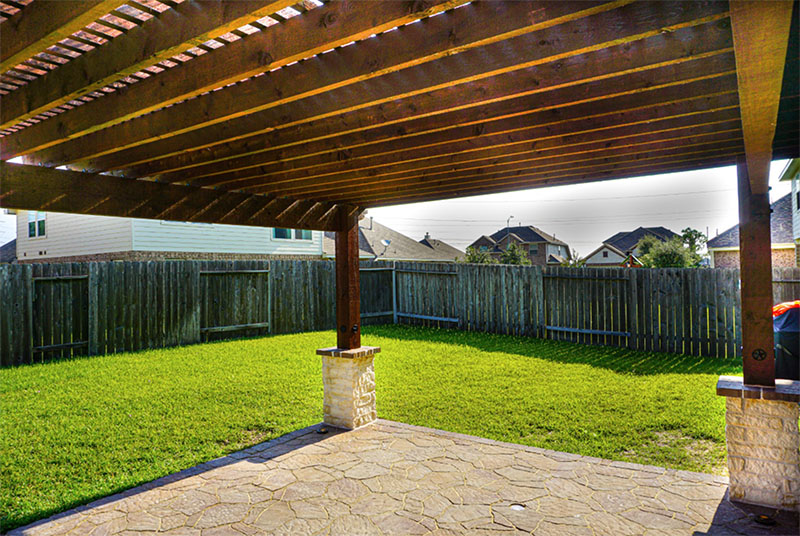 Lattice-covered patio overlooks a green lawn enclosed by a wooden fence; houses in the distance.