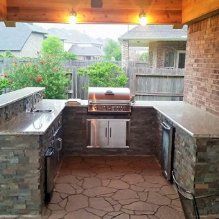 Outdoor kitchen with stone counters, stainless steel grill, and refrigerator under a wooden beam ceiling.