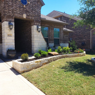 Stone-walled flower bed in front of a brick house with green lawn, sunny day.