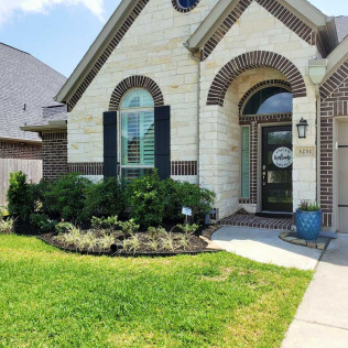 A light-colored stone house with dark arched doorways and black shutters. Green bushes and a blue planter sit in the front.