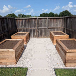 Four wooden raised garden beds with soil, arranged around a stone path in a fenced yard.