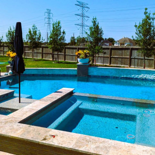Backyard pool with spa, blue water, stone surround, black umbrella, and trees in background.