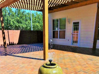 Patio with brick flooring, pergola, fountain, and a building with windows and a door.