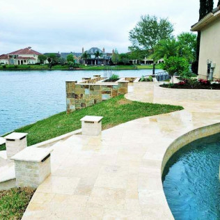 Stone patio next to a pool and lake with houses in the background on a cloudy day.