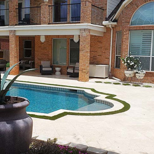 Backyard pool with a house in the background. Brick exterior, white patio, and green artificial turf.
