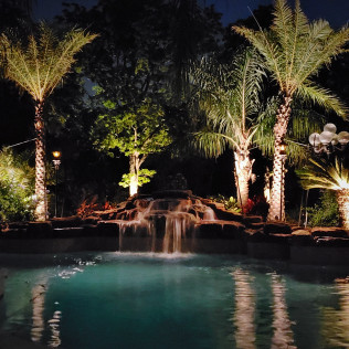 Pool with waterfall at night, lit by landscape lights; palm trees and foliage.