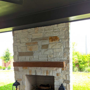 Stone fireplace with wooden mantle under a black overhang, viewed outdoors.