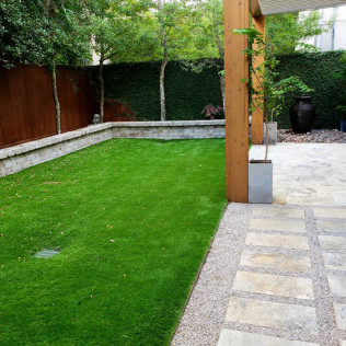 Green lawn bordered by stone patio and gravel, wooden beams, lush green wall.