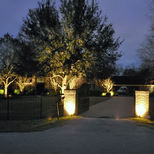 House with outdoor lights illuminating trees, gate, and driveway at dusk.