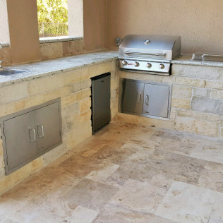 Outdoor kitchen with stainless steel grill, cabinets, and a refrigerator built into stone countertops and flooring.