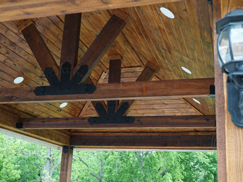 Wooden gazebo ceiling with dark beams, black metal brackets, and recessed lights.