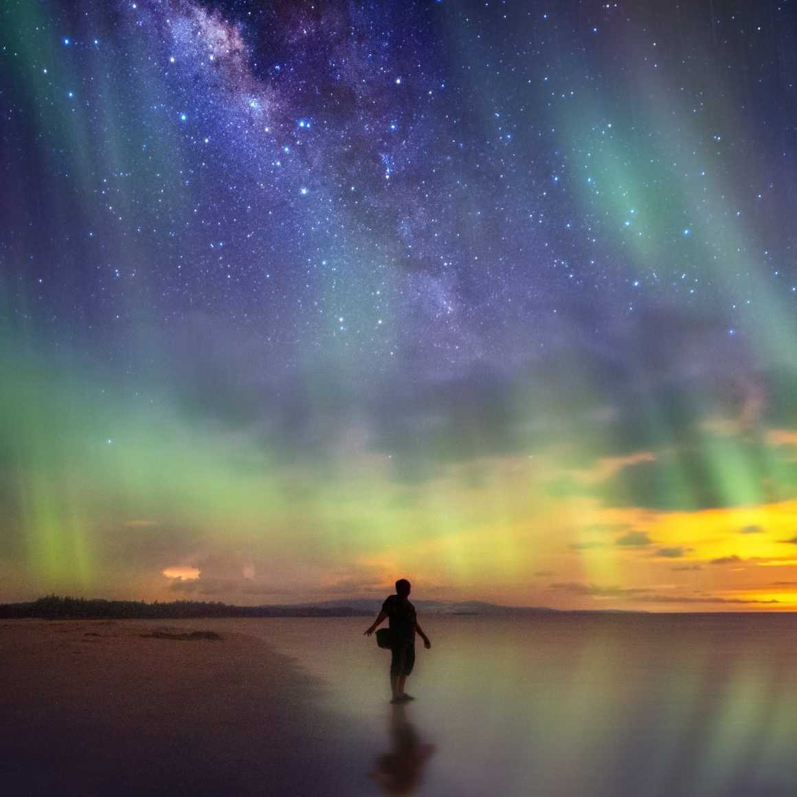 A person is standing on a beach under the aurora borealis.