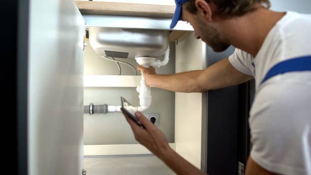 A man is fixing a sink in a kitchen while holding a cell phone. Northglenn, Colorado accountable home services
