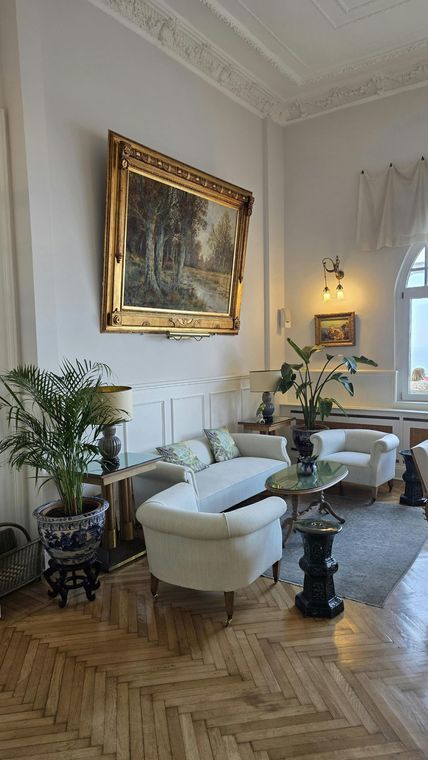 Elegant living room with gold-framed art, white chairs, plants, and a patterned rug on herringbone wood floors