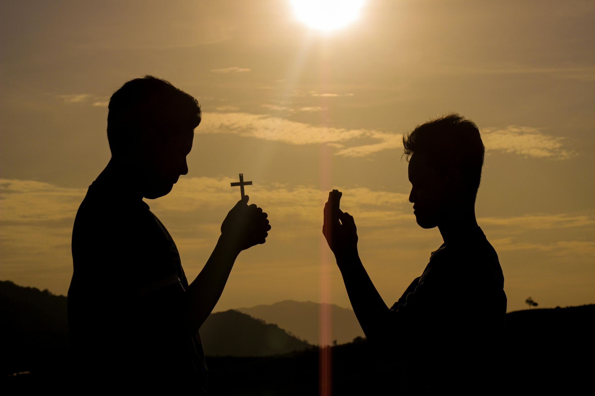 Two silhouetted people facing each other at sunset, one holding up a small cross and the other a phone.