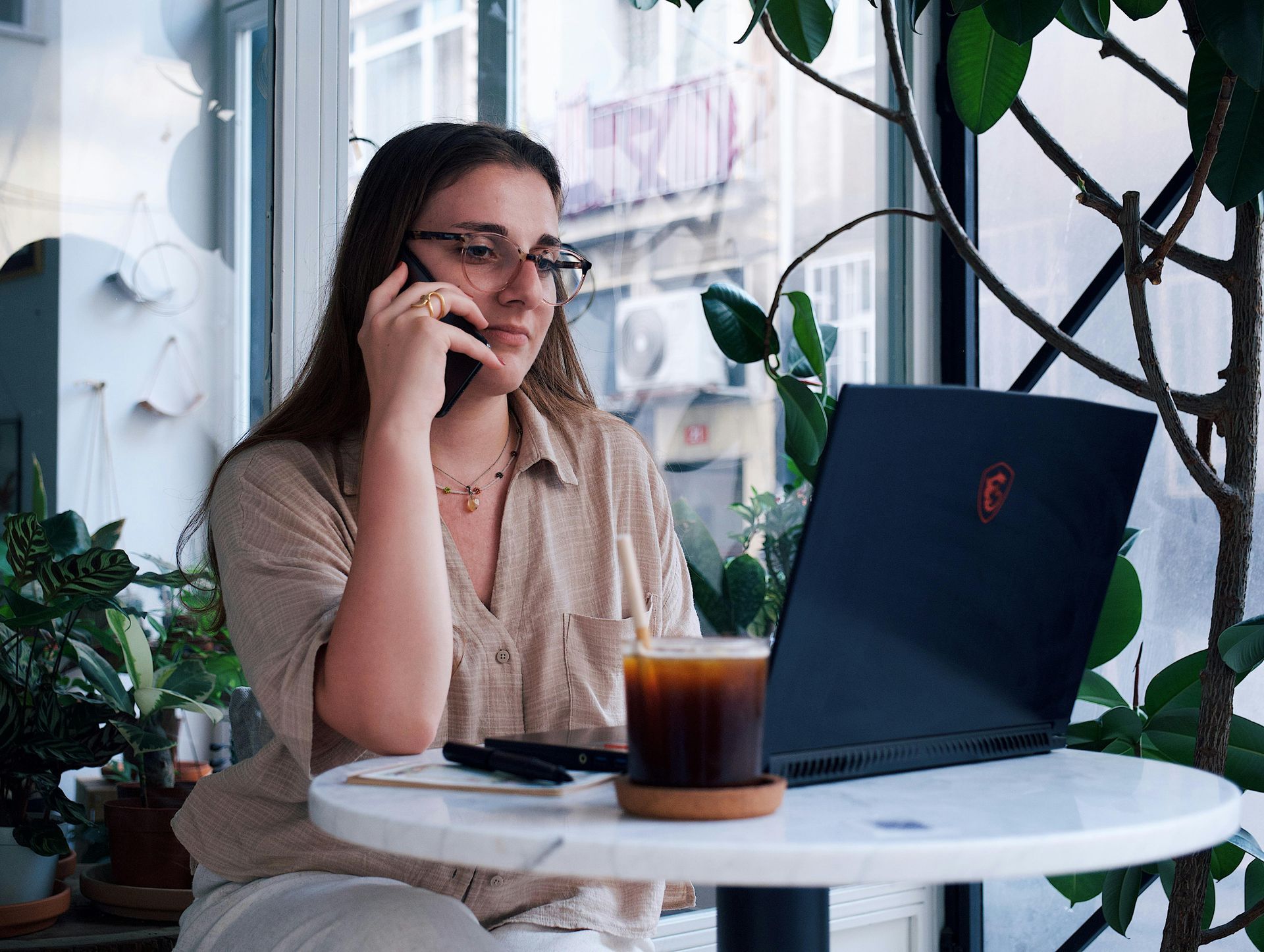 Woman on phone at a small café table with a laptop and iced drink near a window