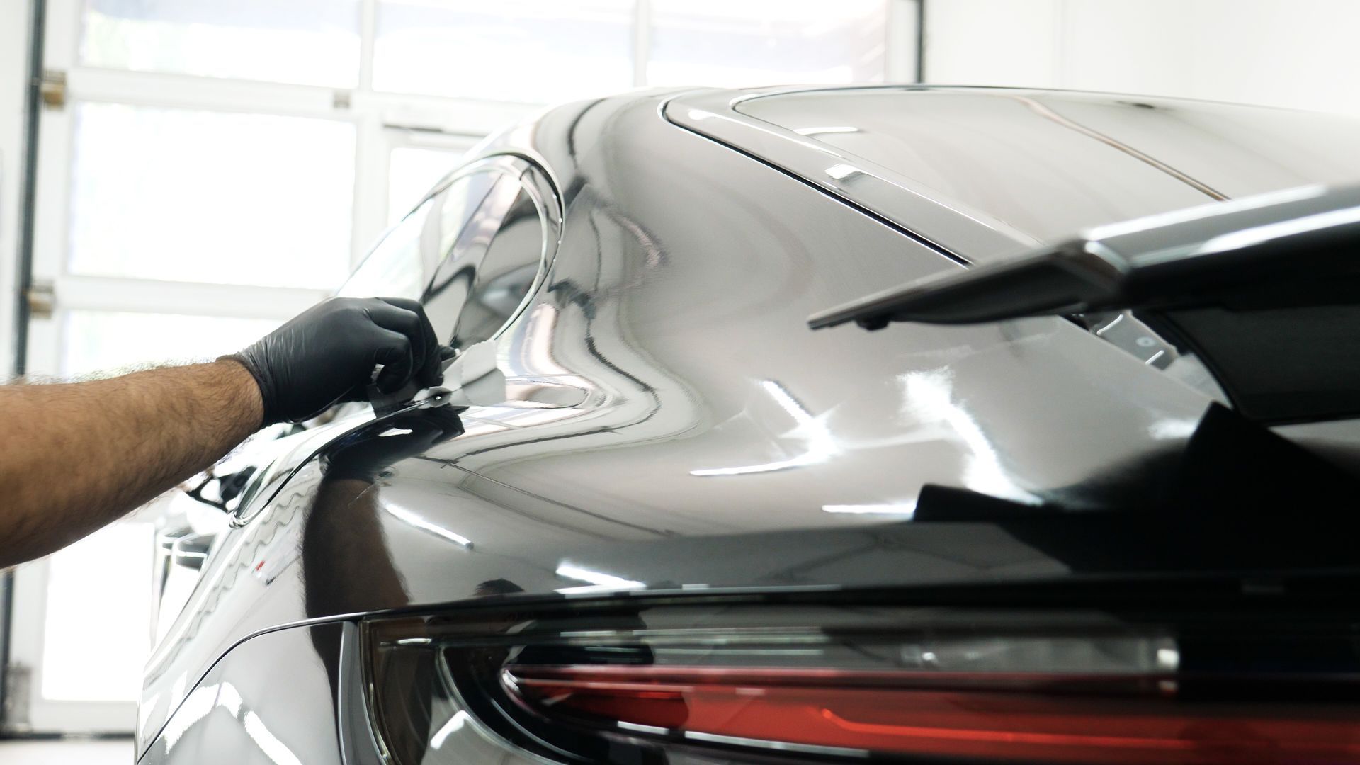 A person in black gloves applying protective film on the rear of a black sports car.