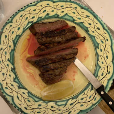 Fettine di bistecca grigliate su un piatto decorativo con uno spicchio di limone e un coltello.