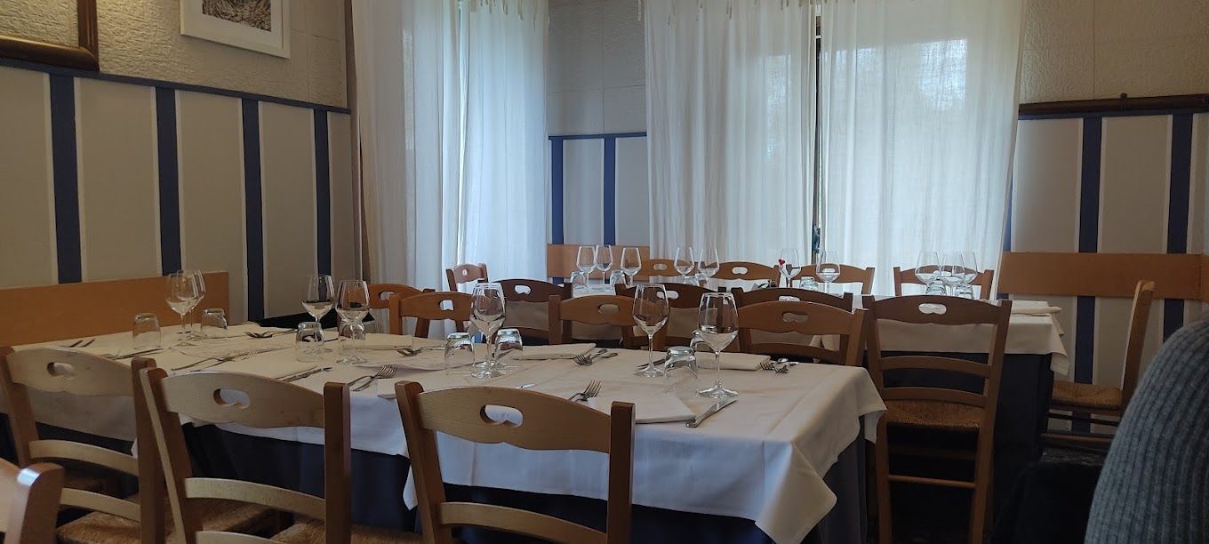 Sala da pranzo con tavoli apparecchiati, sedie in legno e tende trasparenti che filtrano la luce del giorno.