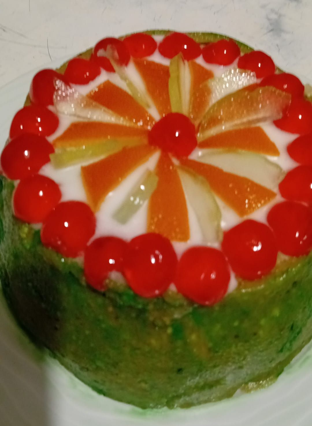 Torta rotonda decorata con frutti rossi, fette di arancia e una ciliegia al centro, con i bordi verdi.