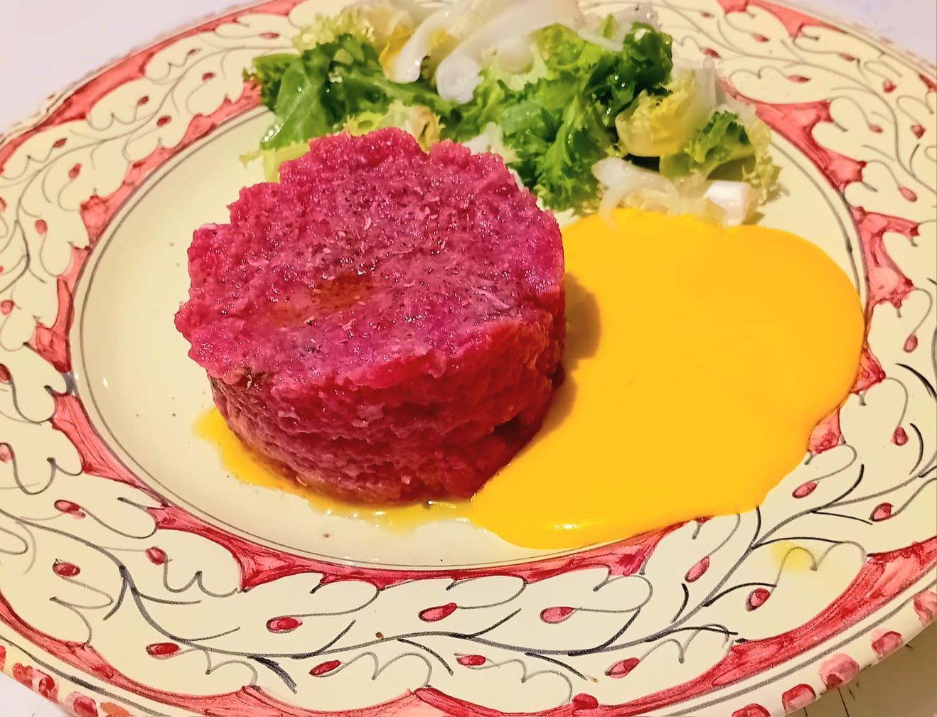 Tartare di barbabietola con salsa gialla e verdure servita su un piatto decorato con motivi floreali.