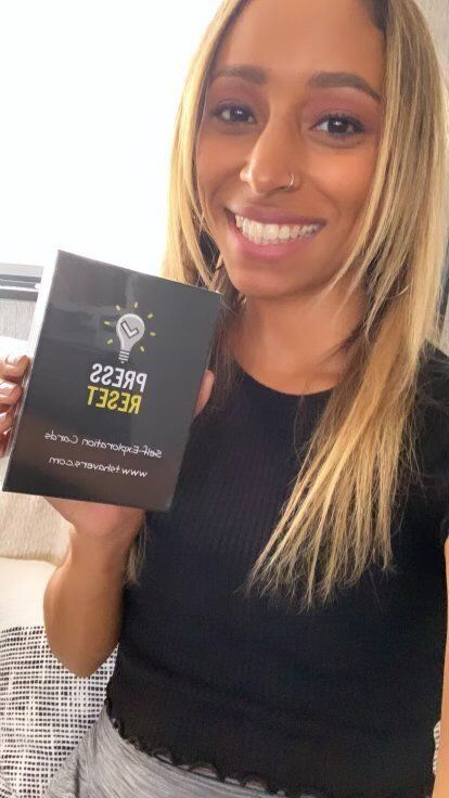 A beautiful young woman with long hair smiles and holds her new Press Reset affirmation card deck.
